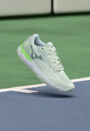 Bílo-zelená sportovní bota se šněrováním, vznášející se nad modro-zeleným povrchem tenisového kurtu u čáry u sítě.