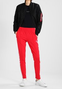 Černá bomber bunda, červené tepláky s bílými bočními pruhy, v kombinaci s bílými teniskami. Černé tričko s logem pod bundou.