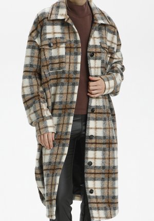 Long manteau à carreaux crème, marron et gris porté sur un col roulé marron et un pantalon noir, montré sur une personne debout du cou à mi-cuisse.