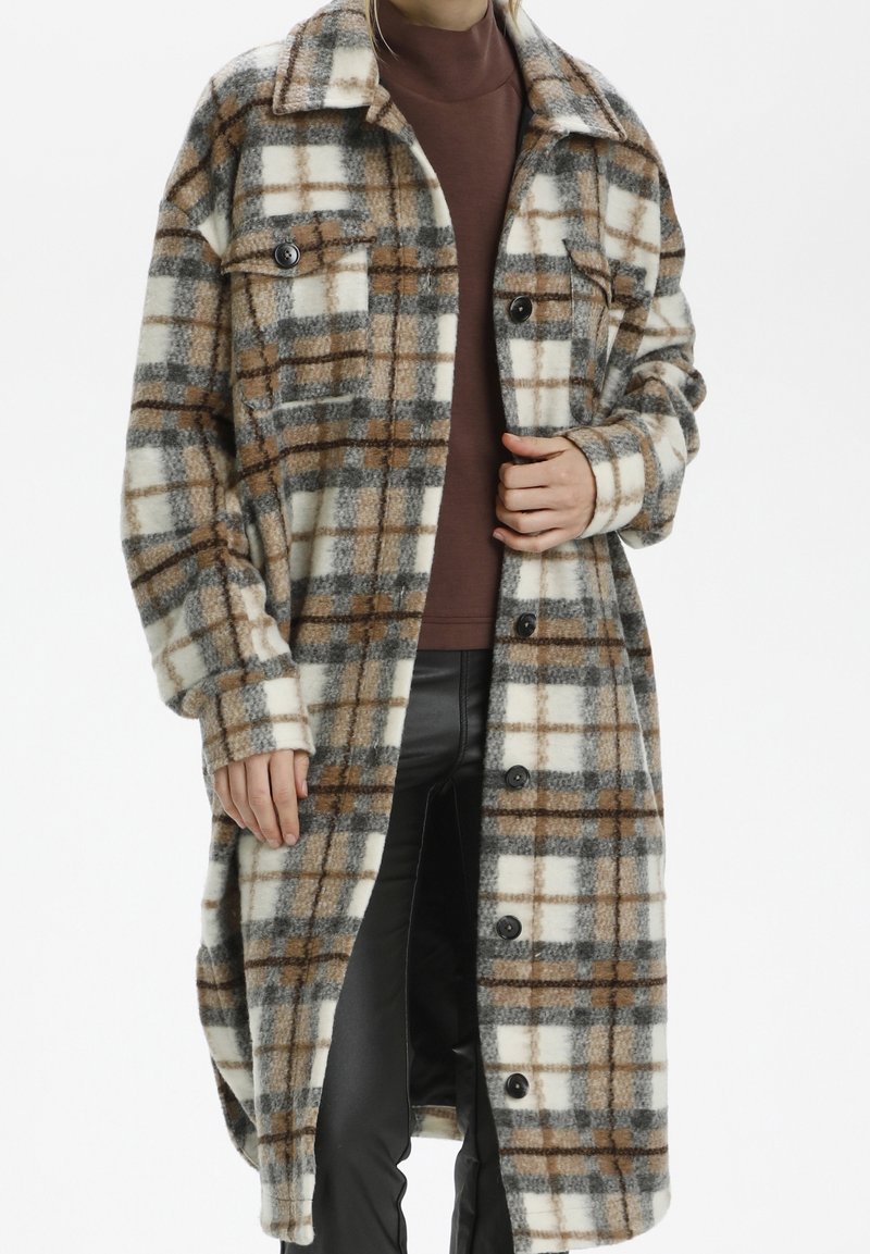 Long manteau à carreaux crème, marron et gris porté sur un col roulé marron et un pantalon noir, montré sur une personne debout du cou à mi-cuisse.