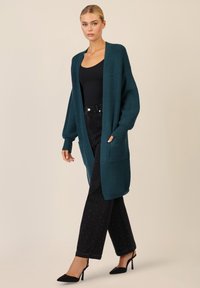 Cardigan lavorato a maglia di colore teal con apertura frontale, maniche lunghe e due tasche frontali, abbinato a pantaloni neri texturizzati e decolleté neri a punta.