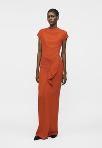 Woman wearing a floor-length, sleeveless burnt orange dress with a ruffled detail on the front, standing against a plain white background.