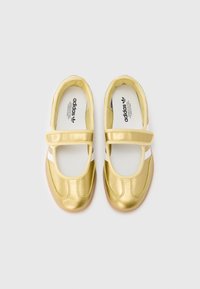 Auksinės Adidas Mary Jane sportbačiai su baltais dryžiais ir lipniomis juostelėmis, žiūrint iš viršaus ant balto fono.