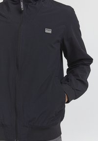 Giacca nera con zip e colletto alto, con polsini e bordo elastici. Presenta una piccola toppa con logo sul lato sinistro superiore. Tessuto leggero.