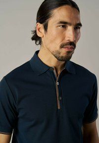 Tumed polo-särk klassikalise kaelusega, kahe nupuga avaga ning pruuni aktsenttikandiga. Lühikesed varrukad ja pehme tekstuur.