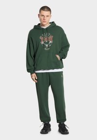 Ciemnozielona bluza z kapturem z graficznym nadrukiem z przodu, kieszenią z przodu oraz dopasowane dresy w tym samym kolorze. Teksturowana bawełniana tkanina.