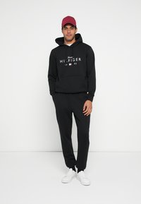 Zwarte hoodie met logo, elastische manchetten en zak, gecombineerd met bijpassende zwarte joggers en witte sneakers. Het model draagt een rode pet.