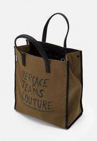 Brązowa torba tote z płótna z czarnymi skórzanymi uchwytami, z nadrukowanym logo "VERSACE JEANS COUTURE" w wyrazistym, ręcznie rysowanym stylu.