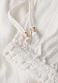 Tela blanca con rayas texturizadas, elástico cinchado con detalles de volantes y dos cordones con acentos metálicos.