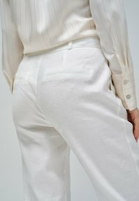 Pantalons en lin blanc avec une texture lisse, dotés de poches arrière et de détails cousus. Porté avec une chemise à rayures fines de couleur claire.