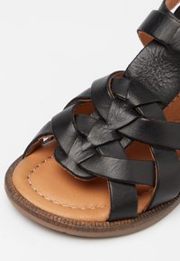 Černá kožená sandál s tkaným svrškem, béžovou koženou stélkou a texturovanou gumovou podrážkou. Obsahuje dekorativní prošívání kolem okraje.