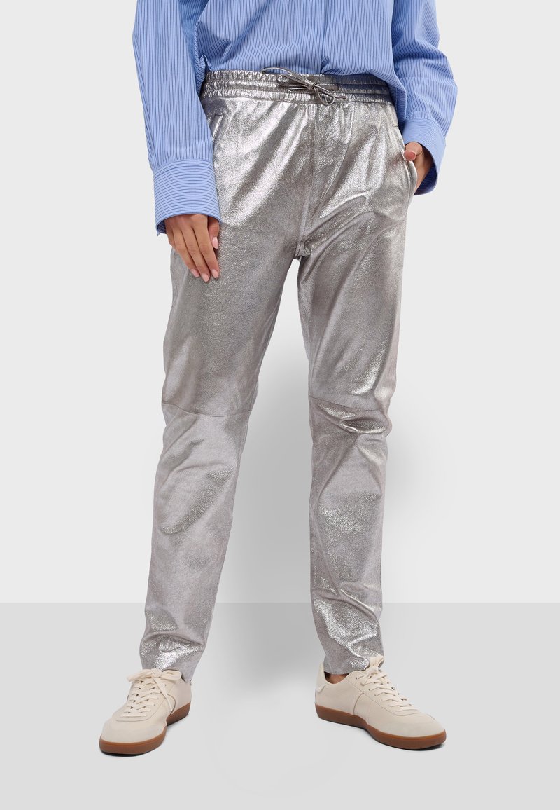 Stříbrné metalické joggingové kalhoty s elastickým pasem a šňůrkou, slim fit, v kombinaci s béžovými teniskami. Hladká, lesklá textura.