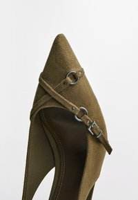 Decolleté in suede marrone con punta affilata, caratterizzato da un cinturino con dettagli in metallo argentato e fibbia sul lato. Fodera interna liscia.