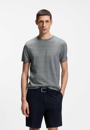 Junger Mann, der ein graues kurzärmeliges T-Shirt und einen schwarzen Gürtel mit dunkelblauen Shorts trägt, steht mit einer Hand in der Tasche vor weißem Hintergrund.