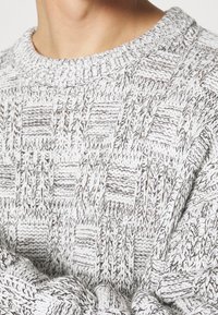 Gros plan d’une personne portant un pull en maille texturée gris clair et blanc avec un motif à carreaux et une encolure ronde.