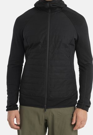 Sort polstret jakke med hætte og fuld lynlås foran båret af en person iført olivengrønne bukser, stående foran en ensfarvet baggrund.