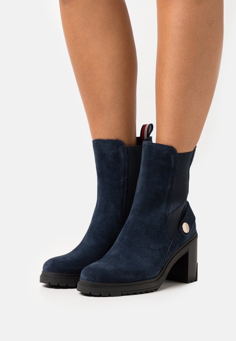 Jambes portant des bottines en daim bleu marine avec des talons blocs, des panneaux latéraux élastiques noirs et une languette rayée à l'arrière.