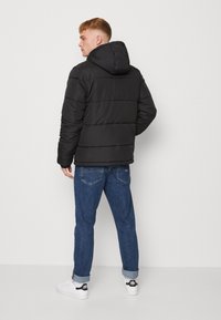 Svart pufferjakke med hette, med horisontale quiltede seksjoner, kombinert med blå denimjeans og hvite joggesko.