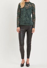 Maglia verde in pizzo a maniche lunghe con motivi floreali, abbinata a leggings in pelle nera e ballerine nere.