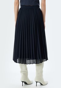 Falda plisada hasta la rodilla en tejido azul marino con un acabado texturizado. Presenta una cinturilla ajustada y una silueta fluida, combinada con botines puntiagudos blancos.