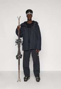 Set de echipament de schi: persoană îmbrăcată într-o jachetă de schi închisă la culoare, pantaloni negri și bocanci de schi, ținând schiurile. Ochelarii de schi și o căciulă completează ținuta de iarnă.