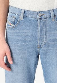 Light blue denim jeans with a straight leg design, featuring orange stitching, a front pocket with a label, and a metal button closure.
