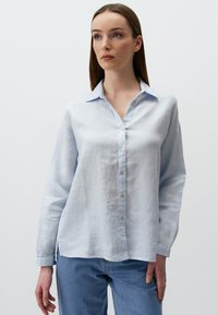 Himmelblaues Leinenhemd mit Knopfleiste, spitzem Kragen und langen Ärmeln. Weiche Textur mit seitlichen Schlitzen am Saum.