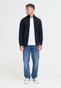 Dunkelgrauer Reißverschluss-Pullover mit hohem Kragen, getragen über einem weißen Hemd, kombiniert mit blauen Jeans und schwarz-weißen Turnschuhen.