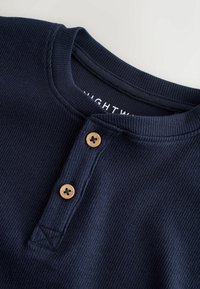 Marineblaues Rippenshirt mit Henley-Ausschnitt, ausgestattet mit zwei Holzknöpfen und einem Etikett mit der Aufschrift "NIGHTW." Weicher, strukturierter Stoff.