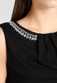 Svart ärmlös topp med en veckad halsringning prydd med klara och silverfärgade detaljer längs kragen för extra stil.