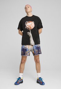Sort oversized t-shirt med grafik og hvidt logo, parret med abstrakte multikolor shorts. Blå sneakers med gule accenter.