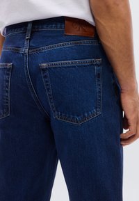 Σκούρο μπλε τζιν παντελόνι με κλασικό σχέδιο, πίσω τσέπες και καφέ δερμάτινο έμπλαστρο με ανάγλυφο λογότυπο της μάρκας στη μέση.