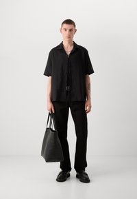 Camisa negra de manga corta con botones, combinada con pantalones negros. El hombre sostiene una bolsa de cuero negra, lleva zapatos negros y un collar de cadena.