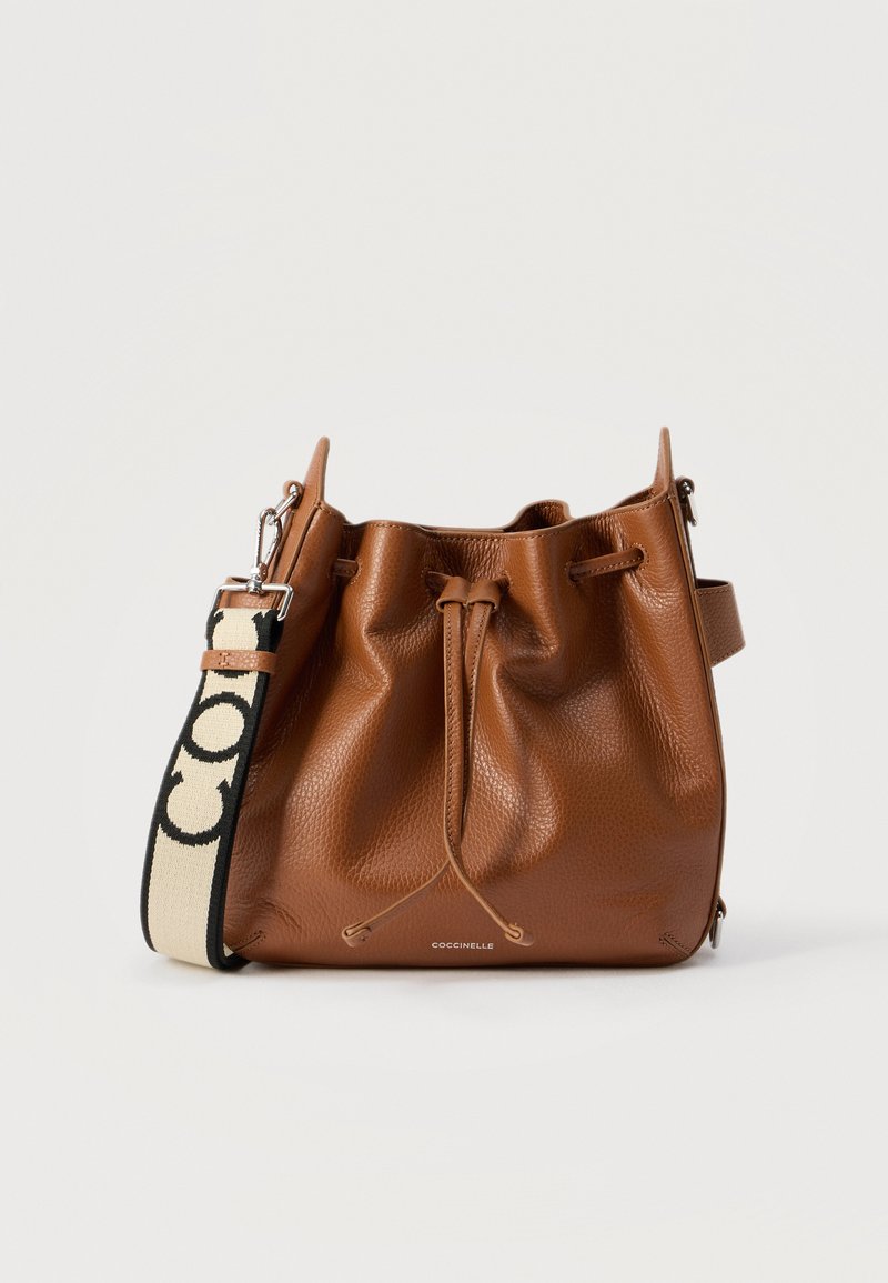 Καφέ δερμάτινη τσάντα τύπου πουγκί με κορδόνι κλεισίματος και μπεζ-μαύρο ιμάντα ώμου με λογότυπο, σε λευκό φόντο.