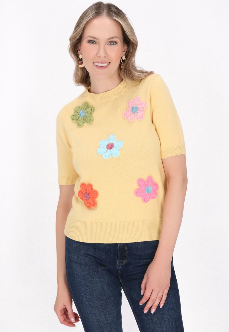 Žlutý pletený svetr s krátkým rukávem a vícebarevnými, texturovanými květinovými aplikacemi. Hladký výstřih a lem, nošený s tmavě modrými džíny.