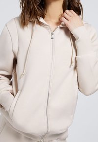 Sudadera con cremallera de color beige claro, con capucha ajustable, puños acanalados y bolsillos laterales. Tela suave y texturizada con un sutil detalle de costura.