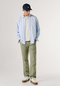 Camisa de botones azul claro sobre una camiseta blanca, pantalones verde oliva, zapatillas blancas con detalles marrones y una gorra azul marino con logo.