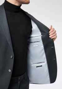 Szary żakiet z gładkiego materiału, z pojedynczym zapięciem na guzik i jasnoniebieską podszewką wewnętrzną. Wiele wewnętrznych kieszeni z etykietami.