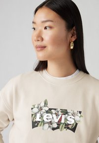 Felpe beige con una grafica floreale che presenta il testo "Levi's" e foglie verdi, colletto rotondo e maniche lunghe. Orecchini a cerchio dorati indossati.