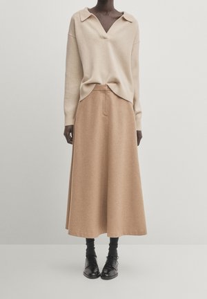 Mannequin portant un pull beige à manches longues et col en V, une jupe midi évasée camel, des chaussettes noires et des chaussures noires à lacets, se tenant devant un fond uni.