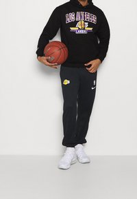 Felpa nera con logo "LOS ANGELES LAKERS" in viola e giallo, abbinata a pantaloni da jogging grigi scuri con emblema dei Lakers e sneakers bianche.