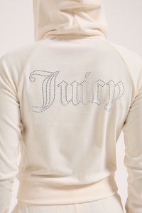 Felpa crema con design aderente, maniche raglan e scritta "Juicy" in strass sul retro. Tessuto morbido con texture liscia.
