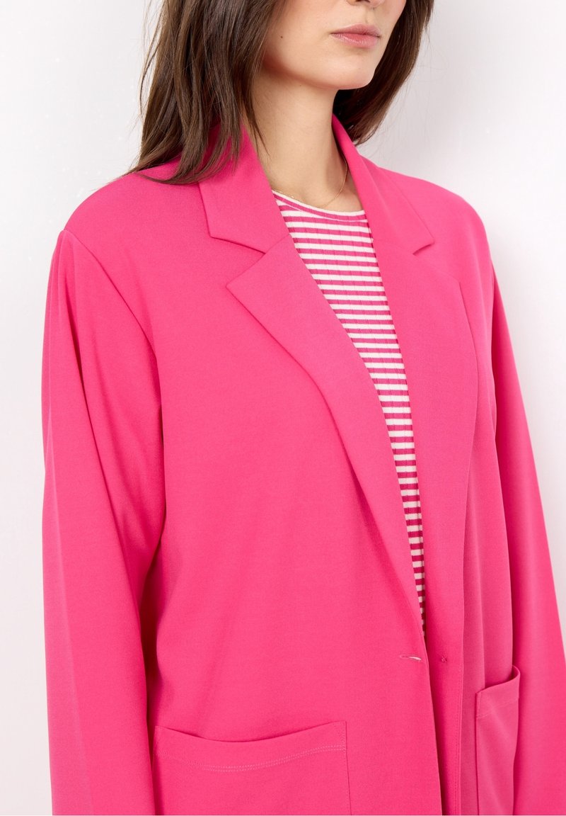 Blazer rosa brillante con rever, due tasche anteriori e tessuto leggero. Indossato sopra una blusa a righe rosse e bianche.