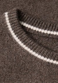 Tessuto di un maglione lavorato a maglia marrone con una texture a coste, caratterizzato da una striscia orizzontale bianca nella zona del collo. Aspetto morbido e testurizzato.