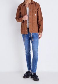 Brun canvasjacka med tryckknappar, bröstficka och dragsko i nederkanten, kombinerad med ljusblå jeans och svarta skor med tofsar.