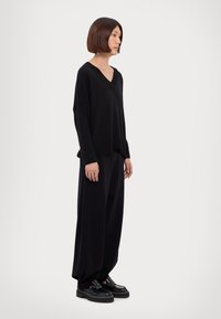 Maglione oversize nero a V con spalle cadenti, abbinato a pantaloni neri larghi e scarpe nere chunky. Texture liscia e design minimalista.