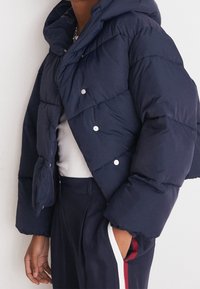 Doudoune courte bleu marine foncé avec finition brillante, dotée d'une capuche et de boutons-pression argentés. Texture lisse avec motif matelassé.