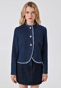 Una donna con capelli biondi e ondulati indossa una giacca di jeans blu scuro, con rifiniture a frange e bottoni, abbinata a una gonna di jeans scura. Sfondo neutro.