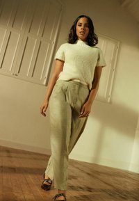 Lys grå, struktureret kortærmet sweater sat sammen med løs, plisseret lys grønne bukser. Modellen står på et trægulv i en afslappet stilling.