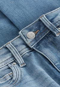 Světle modré džíny z denimu s viditelným knoflíkem a kovovými prvky. Obsahují šité švy a texturovaný povrch. Detailní záběr na pas.